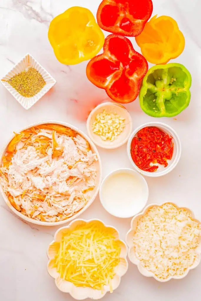 Red, yellow, and orange bell peppers, shredded chicken, cooked rice, sun-dried tomatoes, heavy cream, and fresh Parmesan arranged on a dark surface