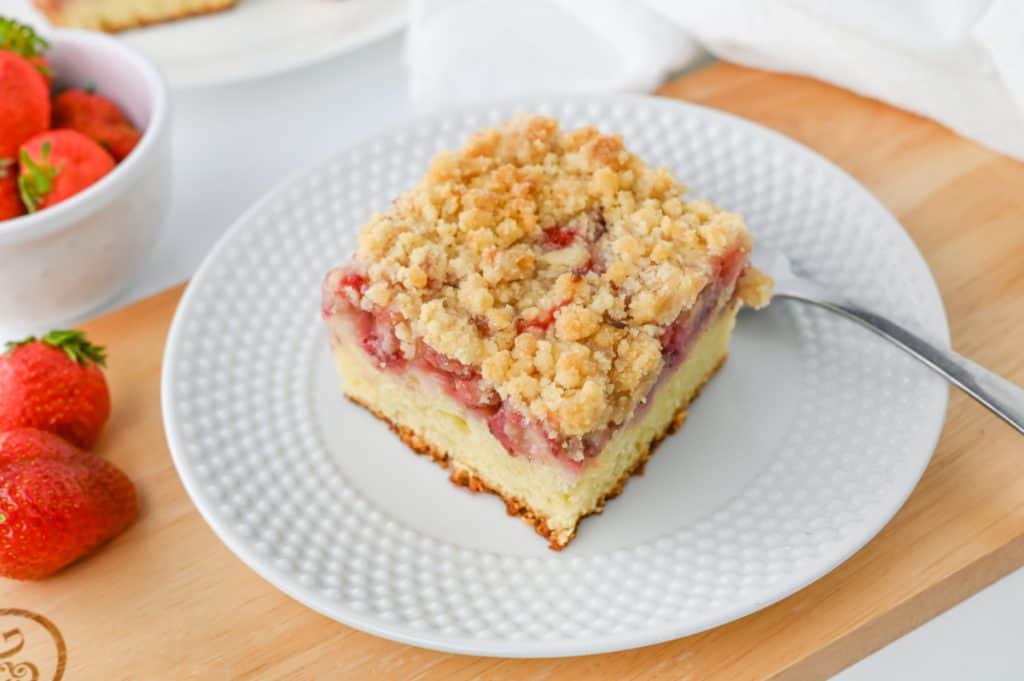 Golden-topped strawberry crumb cake white ceramic plate with bubble designs on a woodne cutting board with buttery crumble and jammy berry layer visible in a cross-section slice
