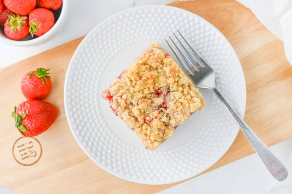 Square slice of strawberry crumb cake on a white plate with a fork, dusted lightly with powdered sugar and served with fresh strawberries
