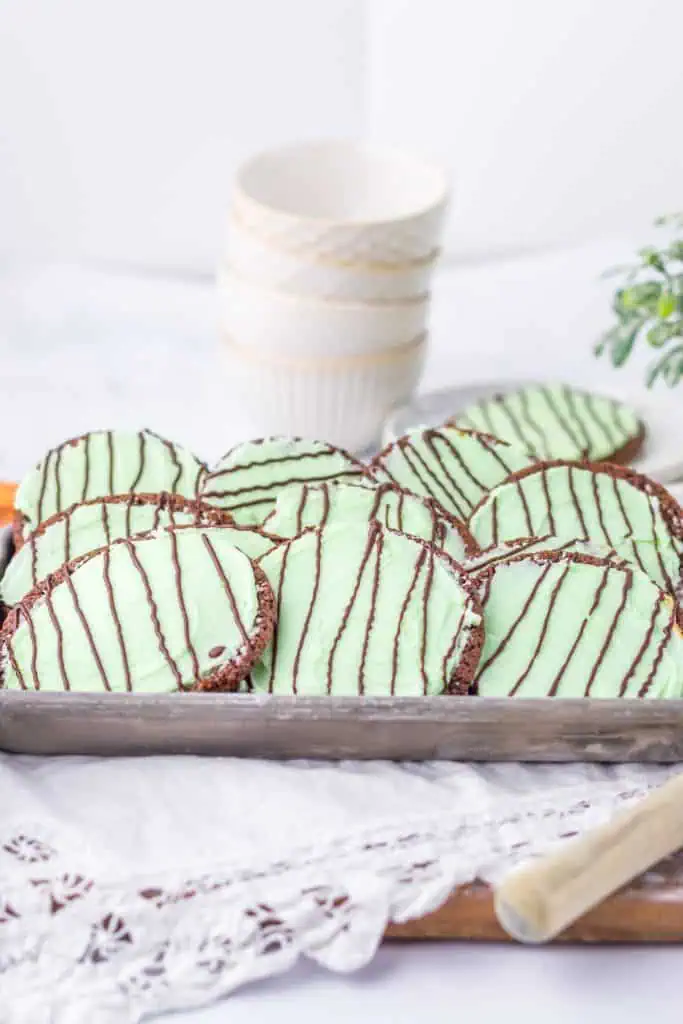Thin mint chocolate cookies on a metal sheet pan beside a glass of cold milk with fresh mint leaves scattered around the base