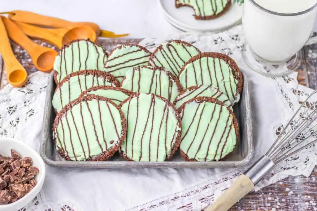 Close-up of a soft thin mint chocolate cookie showing creamy mint green frosting under thin ribbons of hardened chocolate drizzle on a metal pan with a bowl chocolate chips and glass of milk