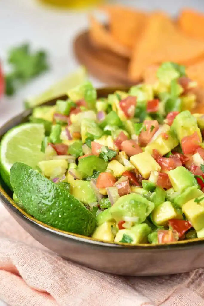 Avocado salsa served in a wide ceramic bowl alongside tortilla chips, a wedge of lime, and a cold drink on a summer outdoor table