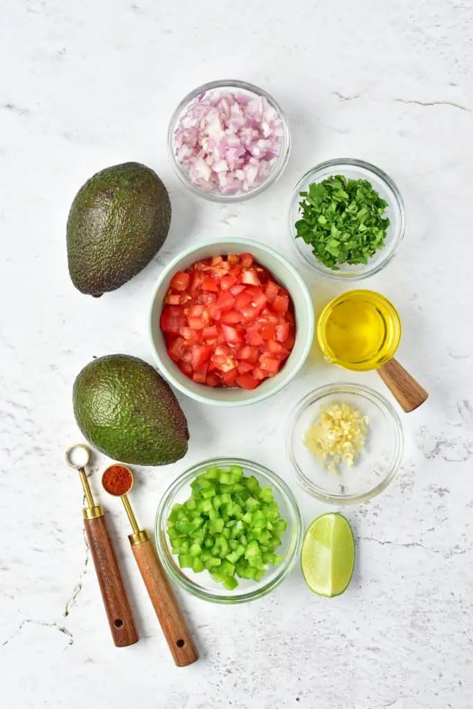 Overhead flatlay of avocado salsa ingredients including halved avocados, red onion, tomato, cilantro, lime, garlic, olive oil, and smoked paprika on a white surface