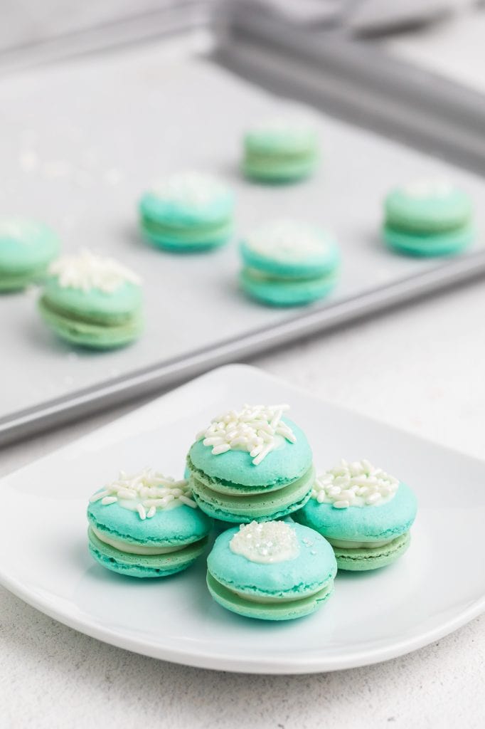 Six blue raspberry macarons arranged in a row on a white marble board, soft natural light, white linen alongside and some stacked on a pile
