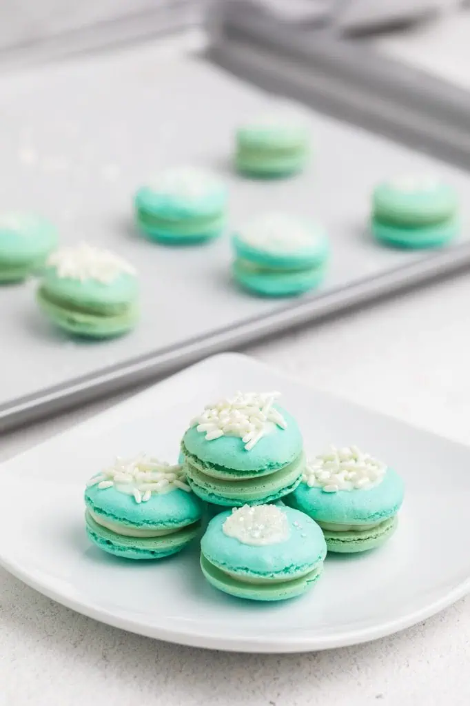 Six blue raspberry macarons arranged in a row on a white marble board, soft natural light, white linen alongside and some stacked on a pile