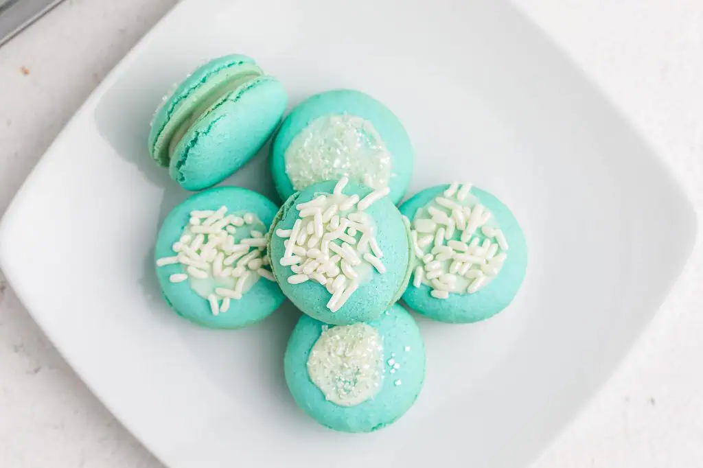Close-up of a single blue raspberry macaron showing the smooth aqua shell, delicate ruffled feet, creamy white chocolate ganache filling, and white sugar crystal topping