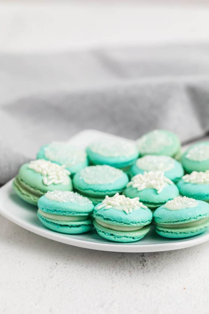 A white square plate piled with soft aqua blue macarons topped with white sprinkles and sanding sugar, filled with white chocolate raspberry ganache
