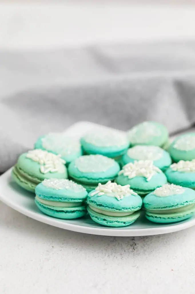 A white square plate piled with soft aqua blue macarons topped with white sprinkles and sanding sugar, filled with white chocolate raspberry ganache