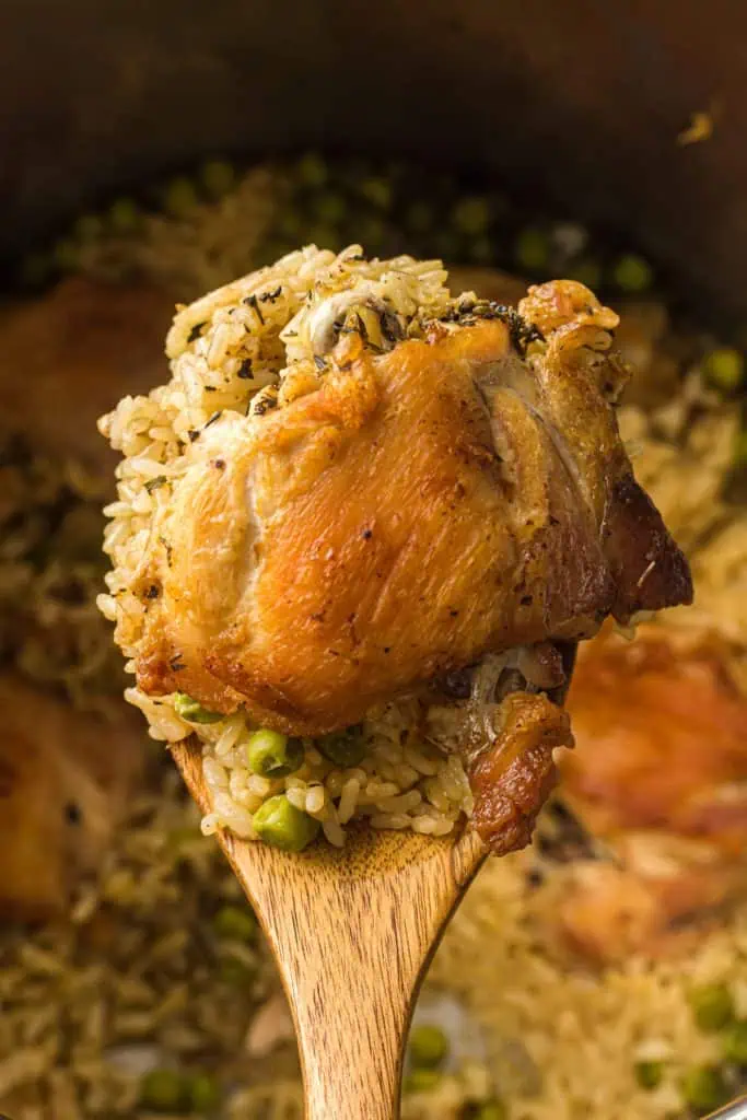 Close-up of a golden-brown seared chicken thigh resting on fluffy seasoned rice with green peas and fresh herbs, coarse black pepper visible on the skin