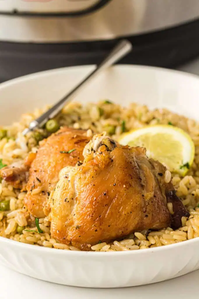A large white bowl of Instant Pot chicken and rice with two golden seared bone-in chicken thighs resting on herb-flecked rice with green peas and a lemon wedge, silver fork alongside, Instant Pot visible in background