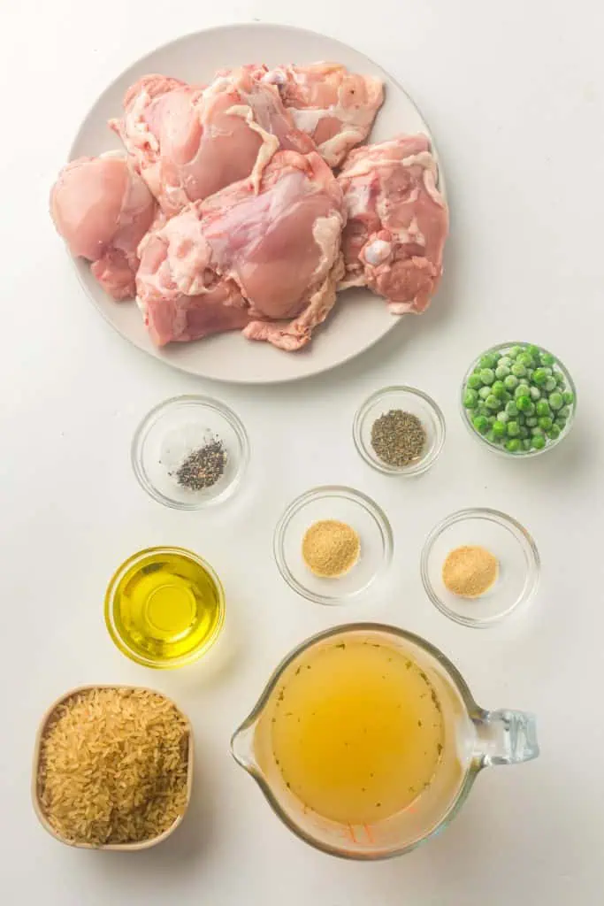 Six bone-in skinless chicken thighs, long grain white rice, chicken broth, frozen peas, olive oil, and spice jars of garlic powder, onion powder, thyme, salt and pepper arranged on a white surface