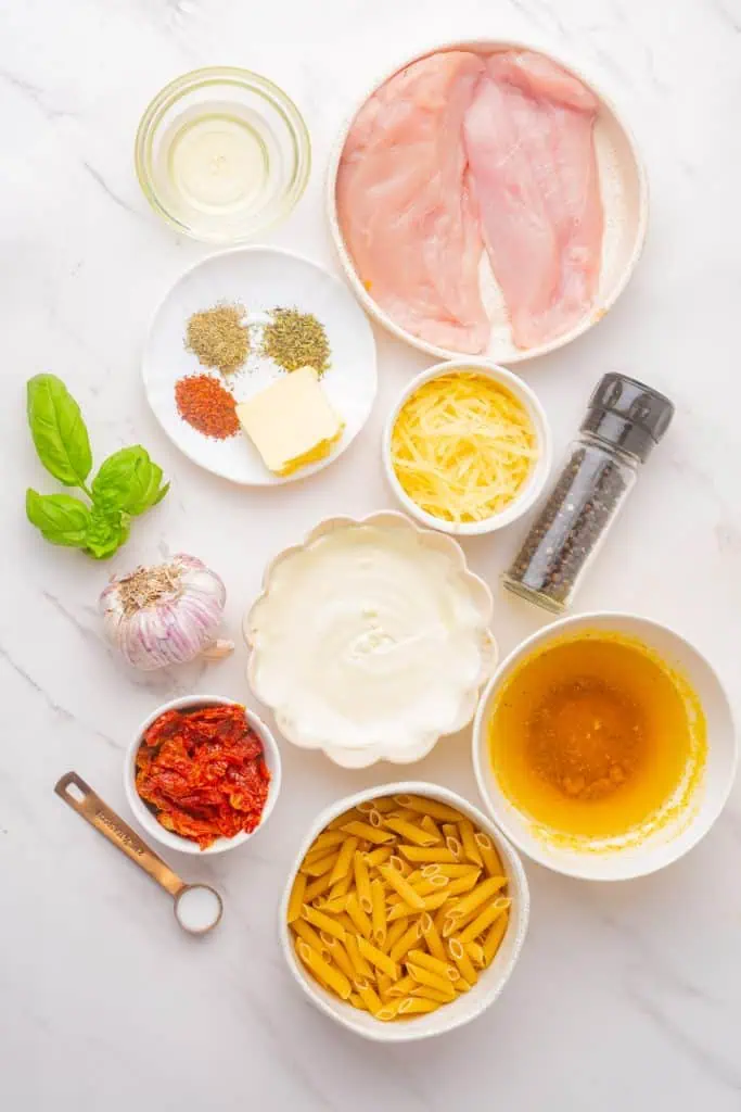 Rigatoni, chicken breast cubes, sun-dried tomatoes, garlic, heavy cream, Parmesan block, and fresh basil arranged on a marble surface