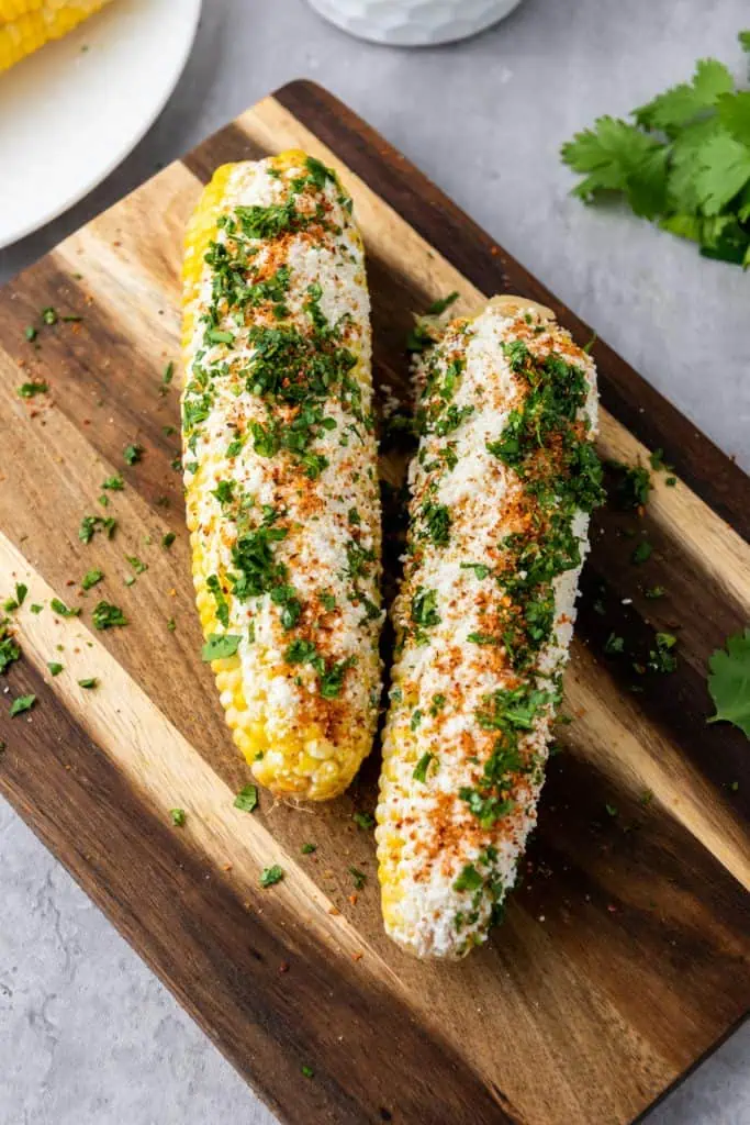 Two fully loaded Mexican elotes on a wooden cutting board, coated in white cotija cheese, bright red tajin, and fresh green cilantro, angled close-up