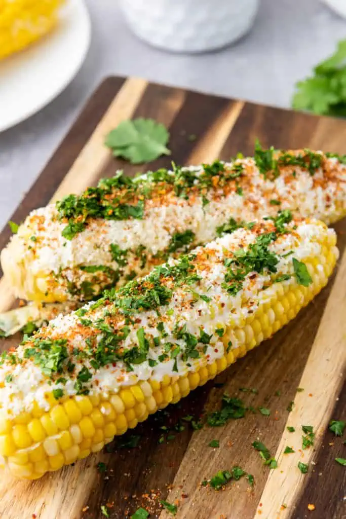 2 fully loaded Mexican elotes on a wooden cutting board, coated in white cotija cheese, bright red tajin, and fresh green cilantro, angled close-up