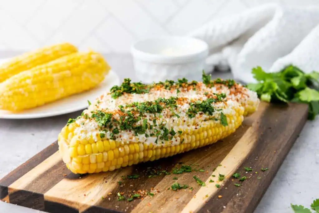 Mexican elotes served on a wooden board with a small jar of crema, lime wedge, and fresh cilantro leaves scattered alongside