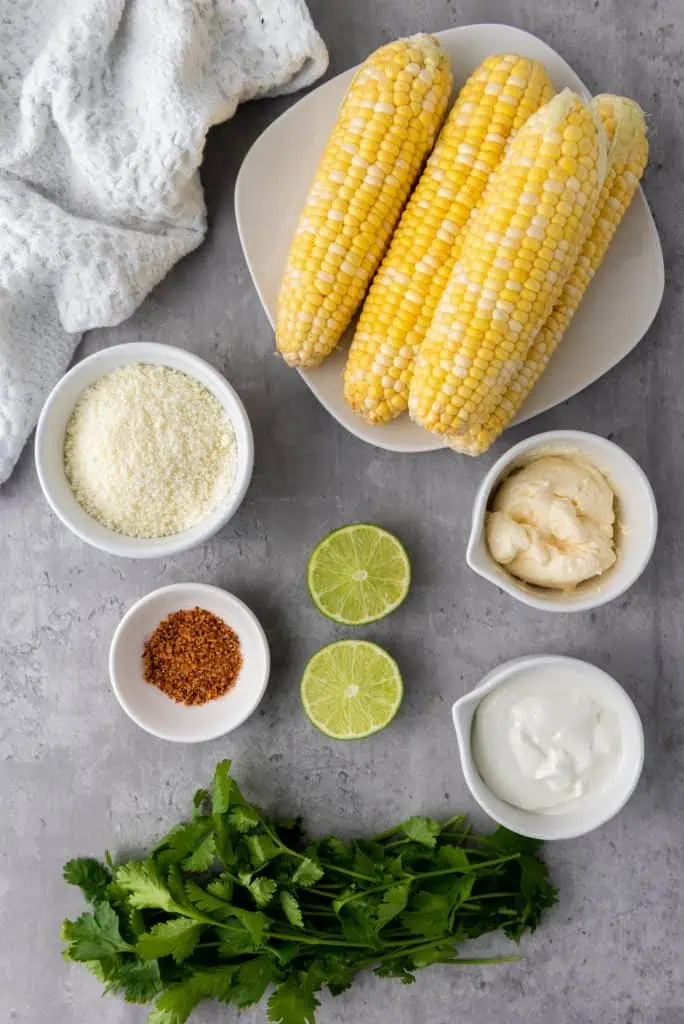 Four ears of corn, container of cotija cheese, Mexican crema, mayonnaise, lime, tajin seasoning, and fresh cilantro arranged on a white surface