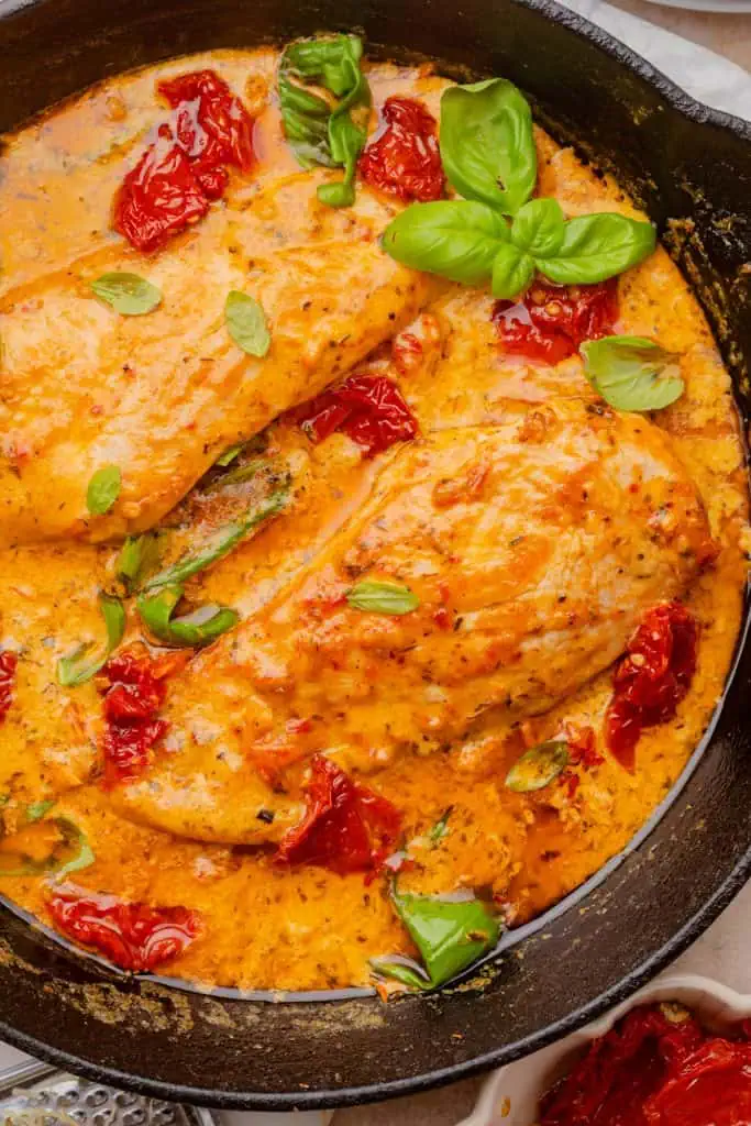 Two golden-seared chicken breasts resting in a pool of creamy sun-dried tomato Parmesan sauce in a cast iron skillet, garnished with fresh basil