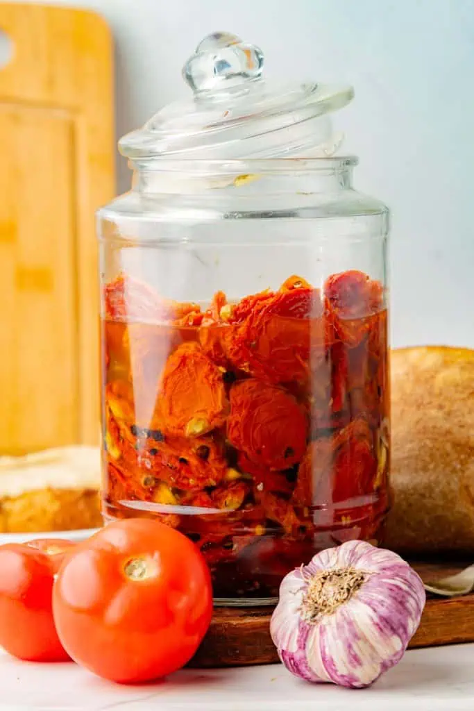 Glass jar filled with golden olive oil and ruby-red sun-dried tomatoes, garlic, and herbs against a light kitchen background