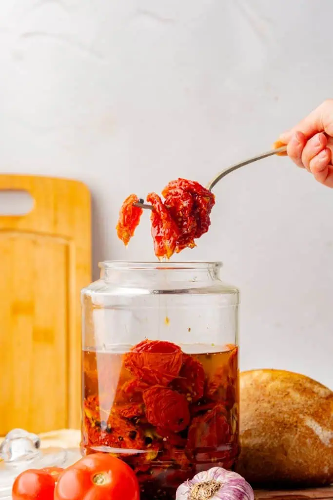 Forkful of homemade sun-dried tomatoes in oil held above an open jar, olive oil dripping back in, natural light kitchen setting
