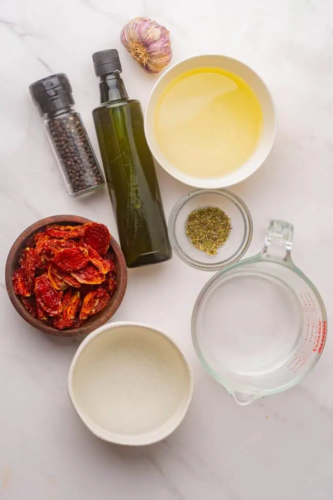 Dry-packed sun-dried tomatoes, garlic cloves, peppercorns, oregano, white vinegar, and olive oil arranged on a white marble surface