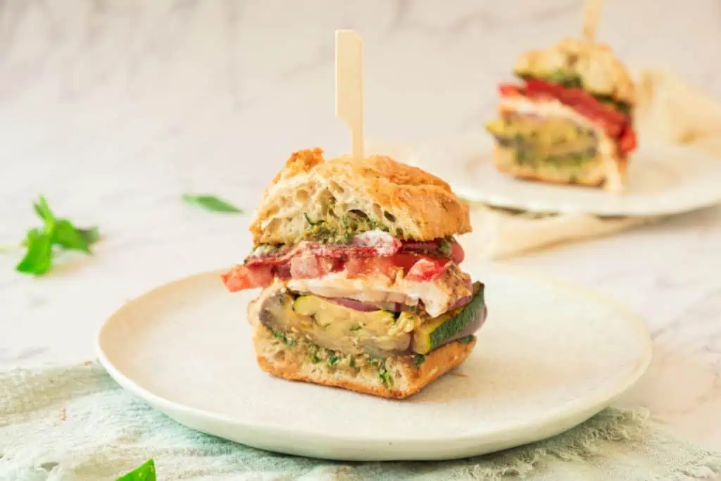Grilled vegetable sandwich half on a white plate with a bamboo pick showing colorful layers of roasted peppers, tomato, burrata, and grilled vegetables