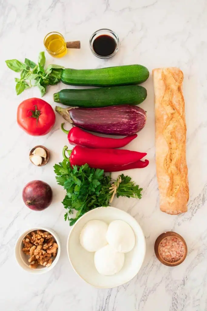 Grilled vegetable sandwich ingredients arranged overhead with zucchini, eggplant, red bell peppers, red onion, tomato, burrata, walnuts, basil, and baguette