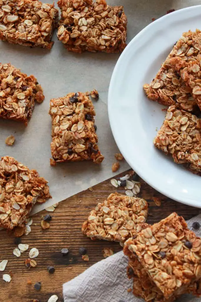 Honey oat granola bars on a white ceramic plate and parchment paper with mini chocolate chips scattered around them and a jar of honey in the background.
