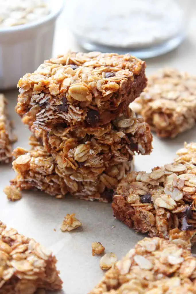 Honey oat granola bars stacked on a wooden cutting board with mini chocolate chips scattered around them and a jar of honey in the background.