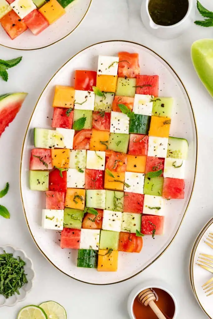 Mosaic melon salad arranged in an alternating pattern on a large white oval serving platter with cubed watermelon, cantaloupe, honeydew, feta, cucumber, and heirloom tomatoes drizzled with honey lime dressing
