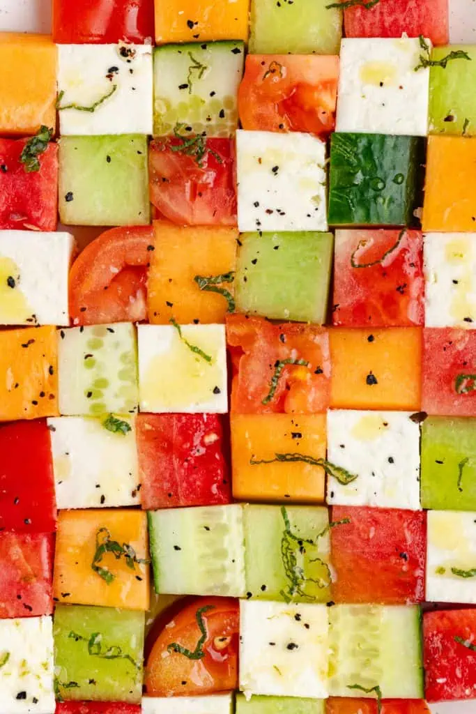 Close-up of mosaic melon salad cubes showing the vibrant red watermelon, orange cantaloupe, green honeydew, white feta, and fresh mint leaves glistening with dressing