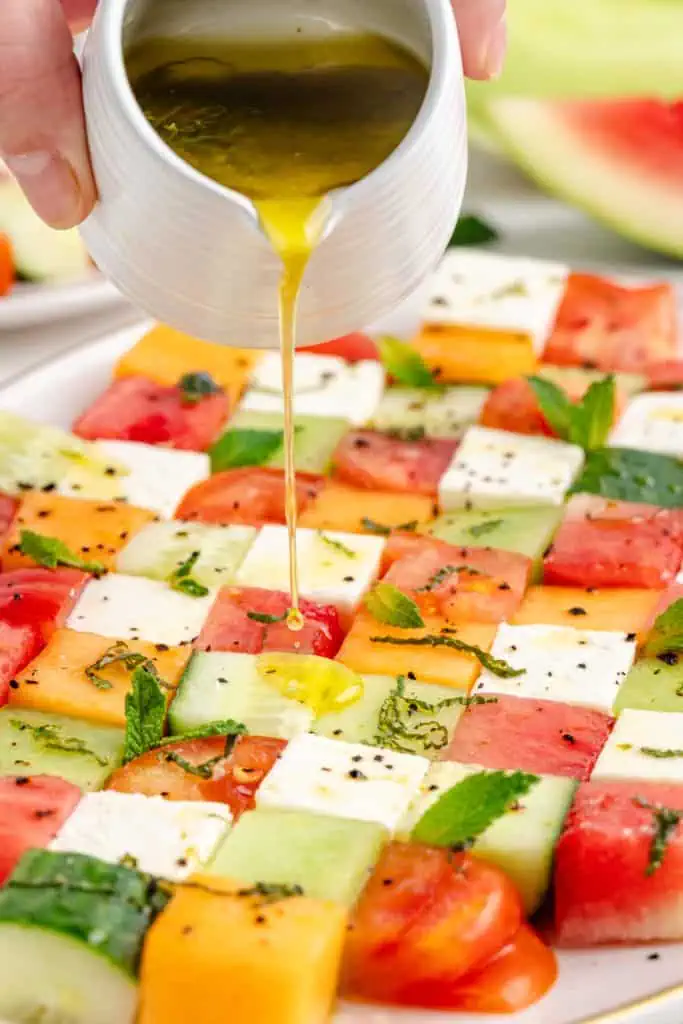 Hand drizzling honey lime mint dressing over arranged cubes of melon, feta, cucumber, and heirloom tomatoes on a white serving platter