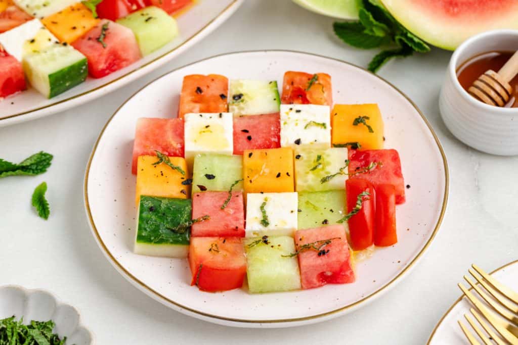 Cubed watermelon, cantaloupe, honeydew, English cucumber, heirloom tomatoes, block feta, and fresh mint arranged on a white marble surface for mosaic melon salad
