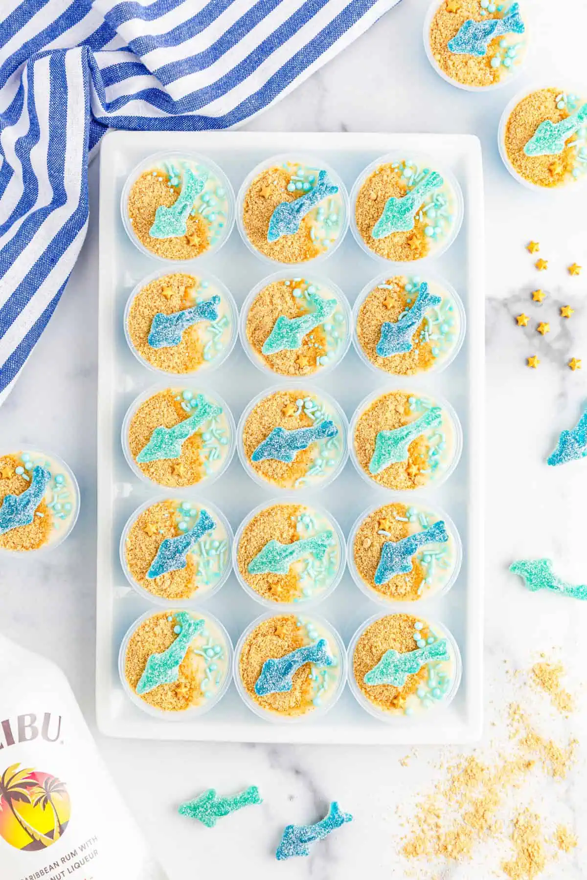Row of layered blue and white shark jello shots topped with graham cracker sand and sour gummy sharks on a white tray