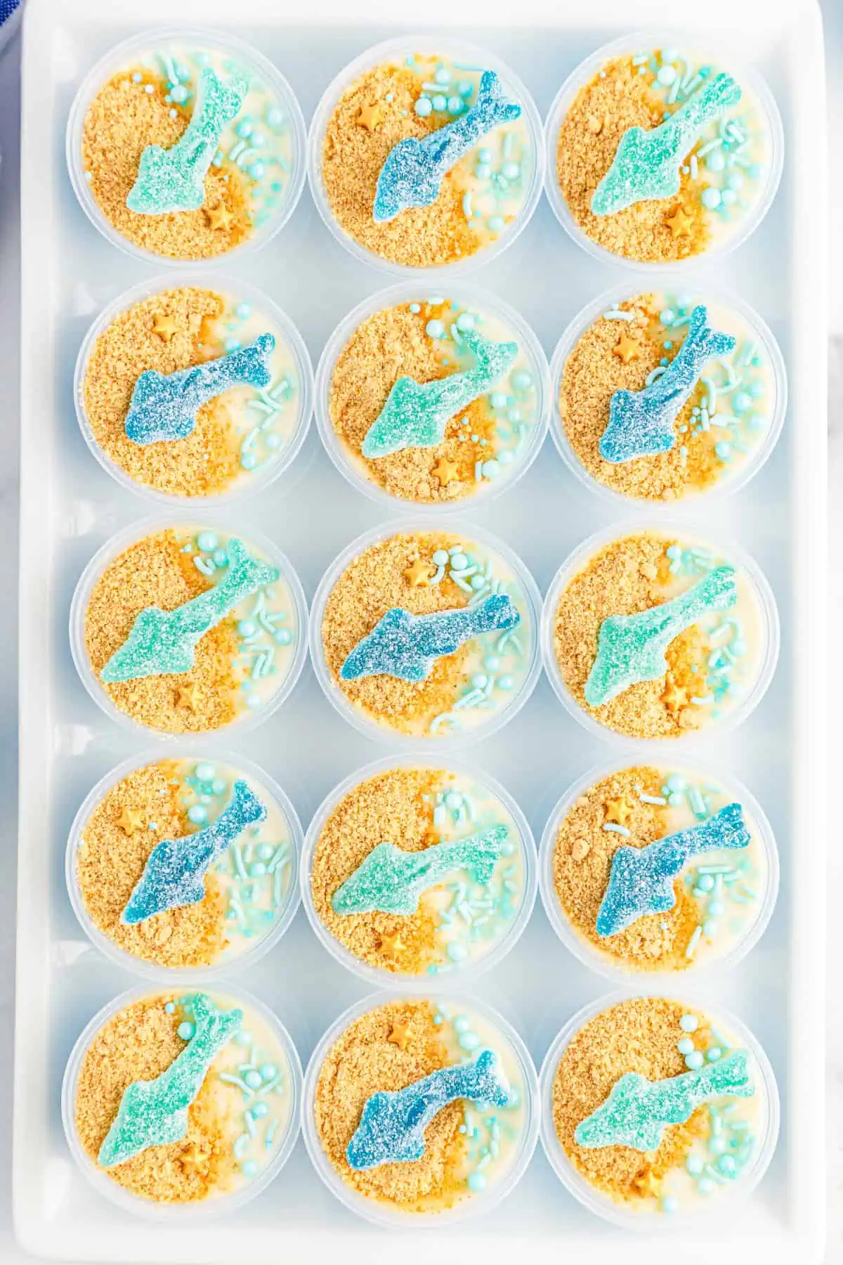 close up view of Rows of layered blue and white shark jello shots topped with graham cracker sand and sour gummy sharks on a white tray