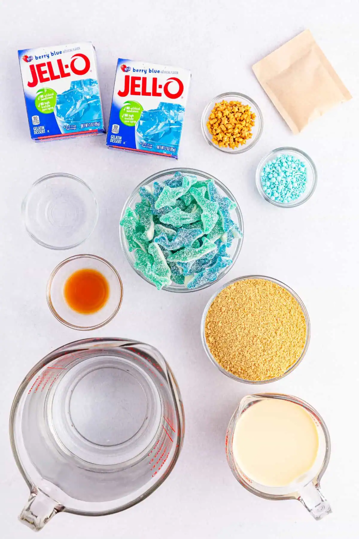 Overhead flatlay of shark jello shot ingredients with blue jello boxes, coconut rum, sweetened condensed milk, graham crackers, and gummy sharks