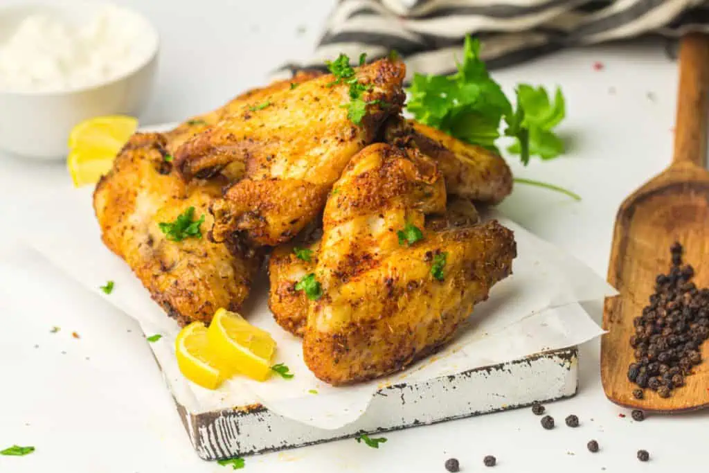 Overhead of golden air fryer chicken wings on a rustic white board with lemon wedges, fresh parsley, and black peppercorns placed in a wooden scoop