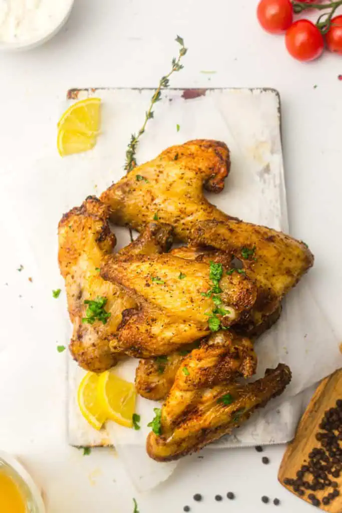 Overhead of golden air fryer chicken wings on a rustic white board with lemon wedges, fresh thyme, and black peppercorns