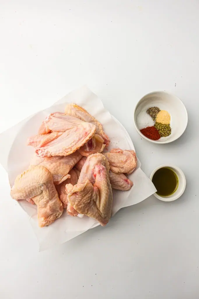 Raw chicken wings on parchment beside small bowls of smoked paprika, garlic powder, herbs, salt, pepper, and avocado oil