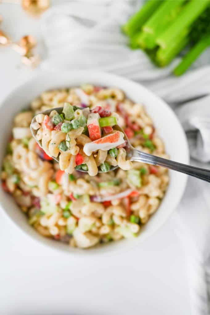 Crab pasta salad served in a white bowl on a bright kitchen counter with fresh celery and wooden serving spoons in the background
