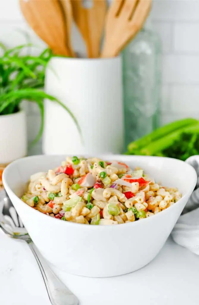 Crab pasta salad in a white bowl with creamy elbow macaroni, chunks of imitation crab, green peas, celery, and red bell pepper topped with fresh chives