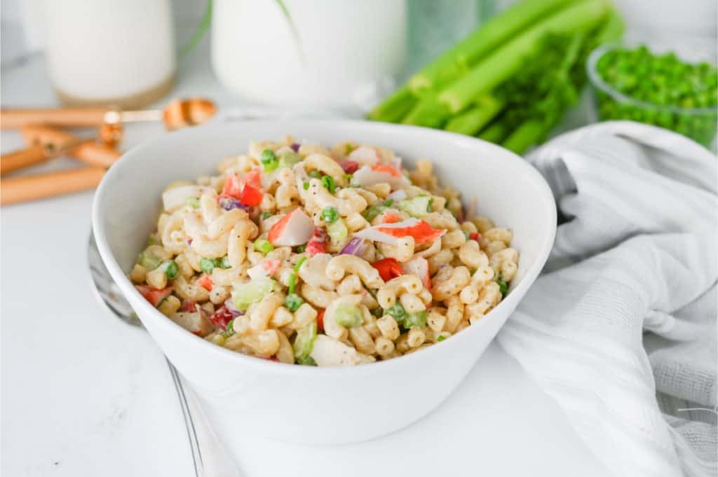 Crab pasta salad served in a white bowl on a bright kitchen counter with fresh celery, peas, and metal serving spoons in the background
