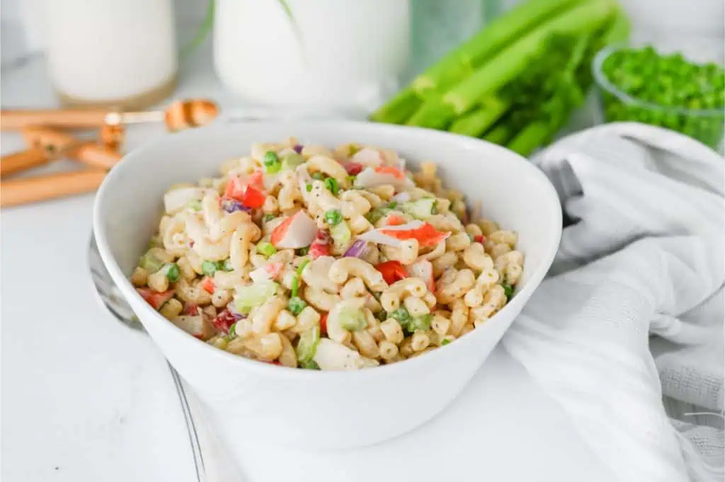 Crab pasta salad served in a white bowl on a bright kitchen counter with fresh celery, peas, and metal serving spoons in the background