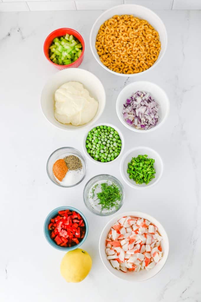 Crab pasta salad ingredients arranged overhead with elbow pasta, imitation crab, mayonnaise, Old Bay, dill, lemon, peas, celery, and red bell pepper
