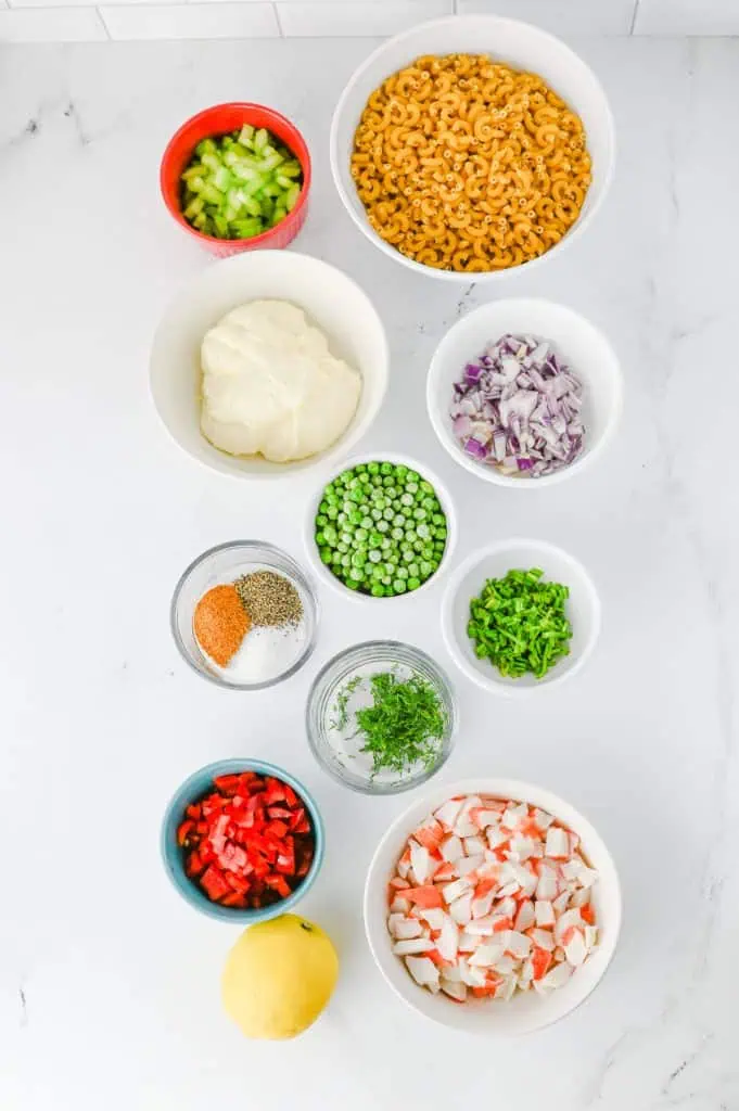 Crab pasta salad ingredients arranged overhead with elbow pasta, imitation crab, mayonnaise, Old Bay, dill, lemon, peas, celery, and red bell pepper
