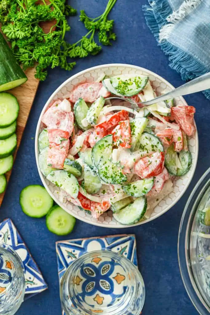 Creamy cucumber tomato salad in a white bowl with thick cucumber half moons, juicy tomato chunks, and sour cream dressing topped with parsley on a dark blue table top and blue napkin