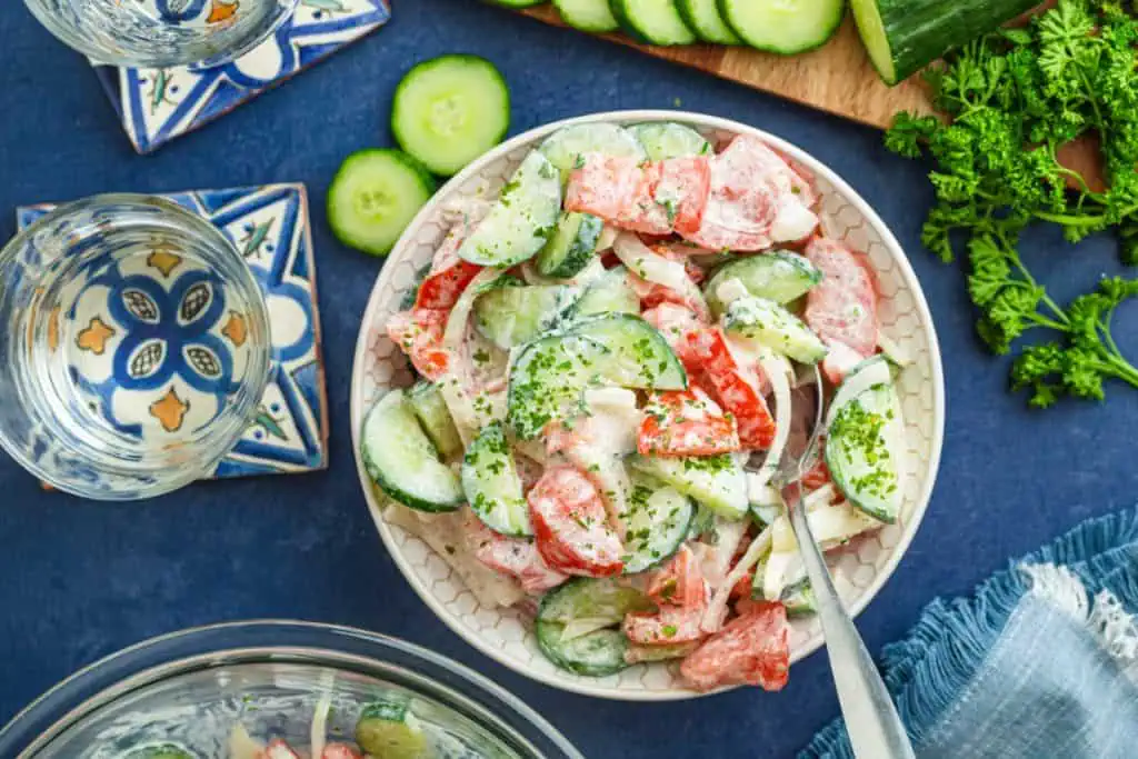Close-up of crunchy cucumber slices and ripe tomato chunks coated in glossy sour cream dressing with cracked black pepper with fresh parsley on a wooden cutting board