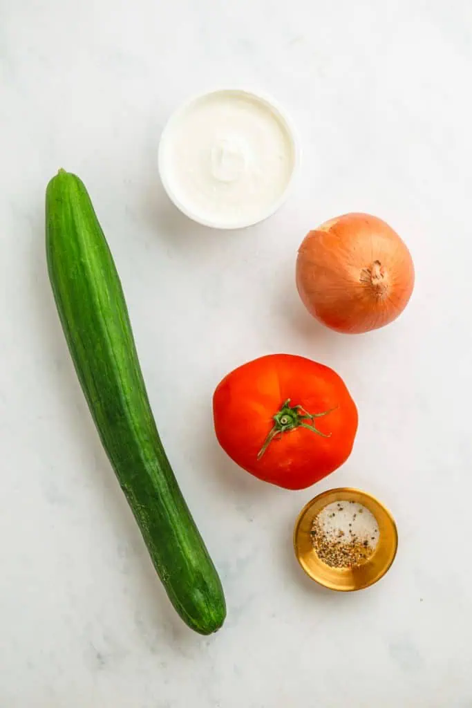 Creamy cucumber tomato salad ingredients arranged overhead with English cucumber, beefsteak tomato, sliced onion, sour cream, and fresh parsley
