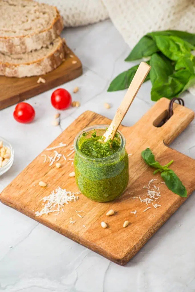 Homemade basil pesto in a glass jar with a wooden spoon, surrounded by grated parmesan, pine nuts, and fresh basil on a wooden cutting board