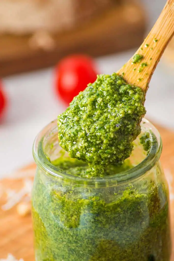 Close-up of vibrant green homemade pesto on a wooden spoon showing flecked texture of blended basil, pine nuts, and parmesan glistening with olive oil