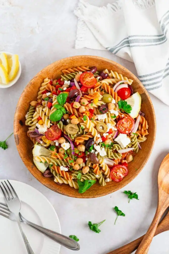 Mediterranean pasta salad in a large wooden brown bowl loaded with feta crumbles, halved olives, colorful peppers, and cherry tomatoes in honey lemon dressing
